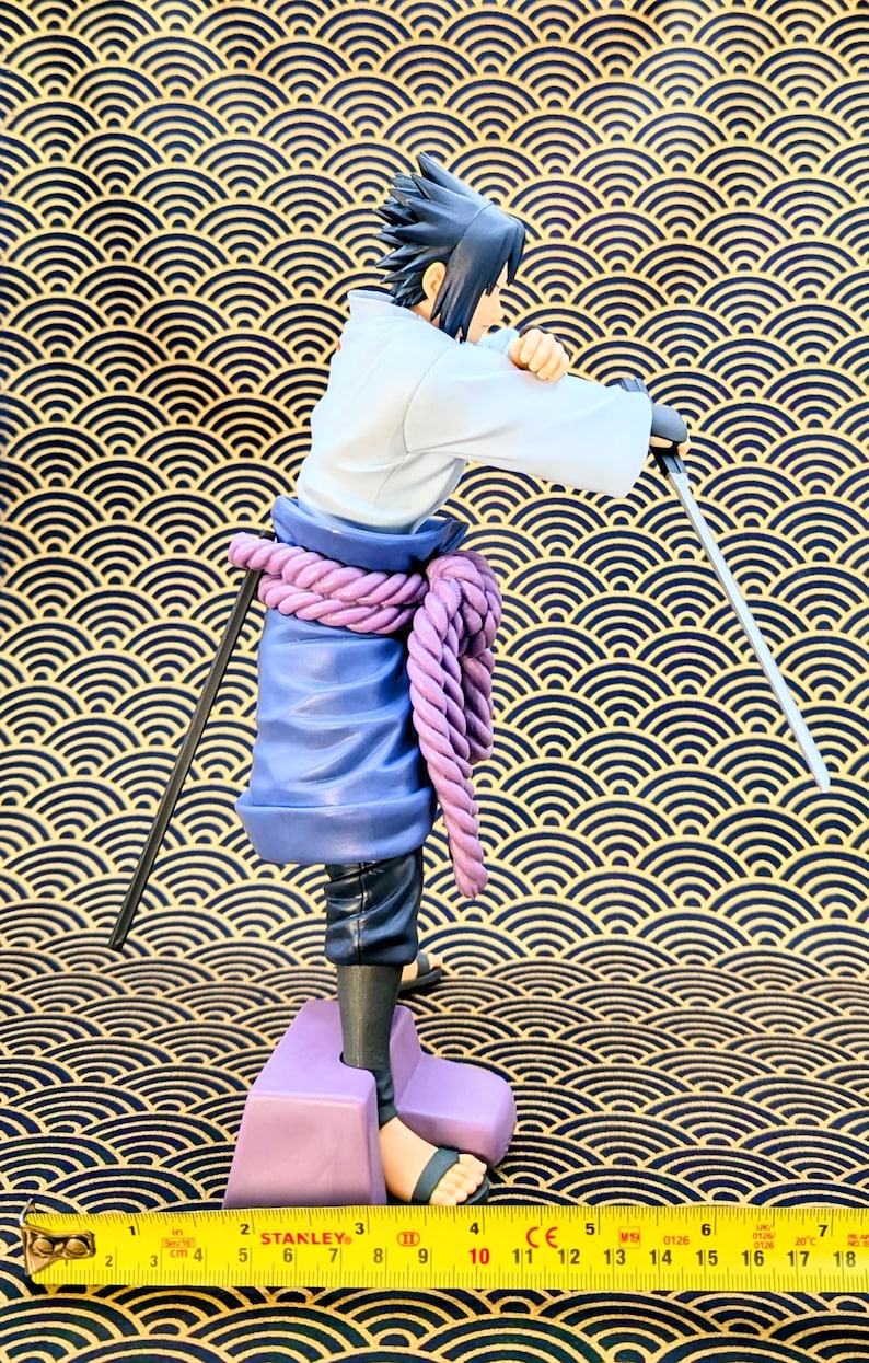 Pode incluir: Uma estatueta de um personagem com cabelo preto espetado, vestindo uma parte superior branca, cal&ccedil;as azuis e um cinto de corda roxa. O personagem est&aacute; segurando uma espada. A estatueta est&aacute; em uma base roxa. O fundo &eacute; um padr&atilde;o de ondas douradas e pretas.