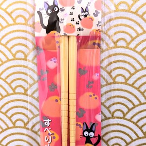 May include: A pair of wooden chopsticks with a pink and white wrapper featuring a black cat character. The wrapper has Japanese text that reads "すべり止め加工" which translates to "non-slip processing". The wrapper also includes the copyright information "&copy; 1989 Eiko Kadono - Studio Ghibli - N".