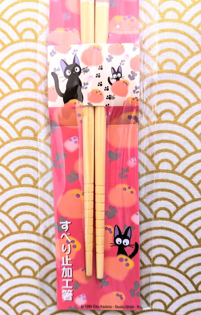 May include: A pair of wooden chopsticks with a pink and white wrapper featuring a black cat character. The wrapper has Japanese text that reads "すべり止め加工" which translates to "non-slip processing". The wrapper also includes the copyright information "&copy; 1989 Eiko Kadono - Studio Ghibli - N".
