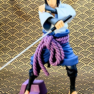 Pode incluir: Figura detalhada de um personagem em uma pose din&acirc;mica, segurando uma espada e dois bast&otilde;es pretos. A figura usa uma roupa branca e azul com um cinto de corda roxa. A figura est&aacute; em p&eacute; em um bloco roxo. Uma fita m&eacute;trica amarela na parte inferior.