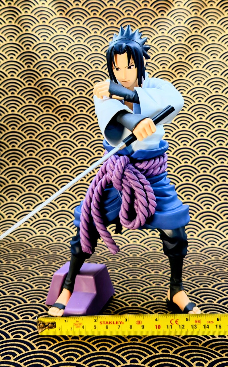 Pode incluir: Figura detalhada de um personagem em uma pose din&acirc;mica, segurando uma espada e dois bast&otilde;es pretos. A figura usa uma roupa branca e azul com um cinto de corda roxa. A figura est&aacute; em p&eacute; em um bloco roxo. Uma fita m&eacute;trica amarela na parte inferior.