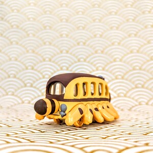 May include: A yellow and brown toy bus with a cartoonish design. The bus has six wheels and a large, round head with a black stripe. The bus is sitting on a white and gold patterned surface.