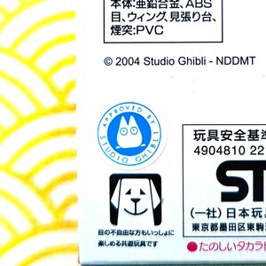 Peut inclure: Gros plan sur une bo&icirc;te blanche avec du texte japonais et des logos Studio Ghibli. La bo&icirc;te pr&eacute;sente un cercle bleu avec un personnage de dessin anim&eacute; et les mots "APPROVED BY STUDIO GHIBLI". En dessous, un carr&eacute; noir avec une illustration de chien.