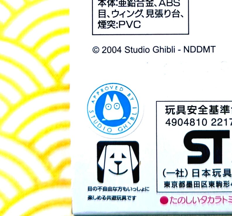 Peut inclure: Gros plan sur une bo&icirc;te blanche avec du texte japonais et des logos Studio Ghibli. La bo&icirc;te pr&eacute;sente un cercle bleu avec un personnage de dessin anim&eacute; et les mots "APPROVED BY STUDIO GHIBLI". En dessous, un carr&eacute; noir avec une illustration de chien.