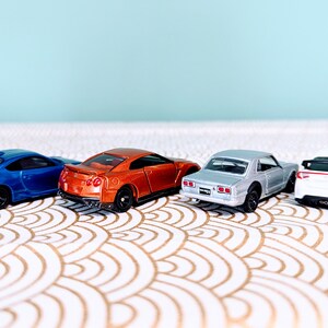 May include: Four miniature toy cars in different colors, including blue, orange, silver, and white, are lined up on a white and gold patterned surface. The cars are facing the same direction, with their rear ends visible.