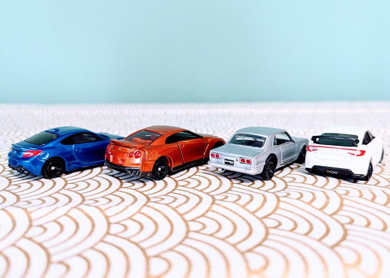 May include: Four miniature toy cars in different colors, including blue, orange, silver, and white, are lined up on a white and gold patterned surface. The cars are facing the same direction, with their rear ends visible.