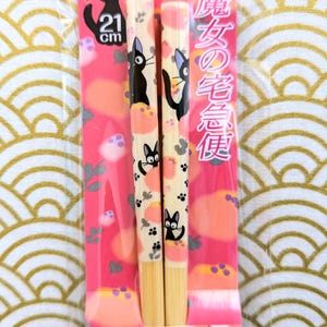 May include: A pair of wooden chopsticks with a pink and white design featuring a black cat with a white face. The chopsticks are 21 cm long and are packaged in a clear plastic bag. The packaging also includes the text "魔女の宅急便" in Japanese.