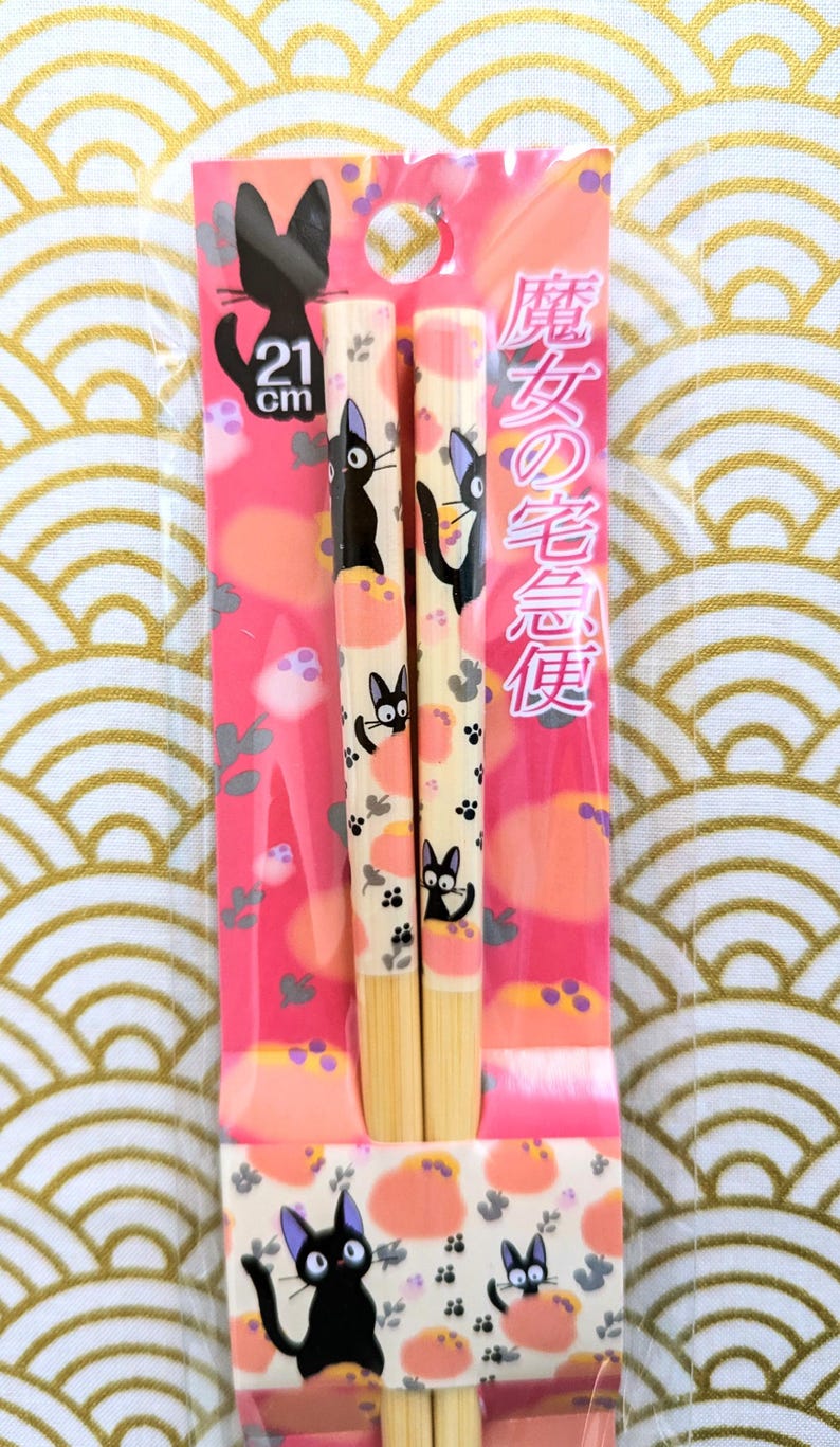 May include: A pair of wooden chopsticks with a pink and white design featuring a black cat with a white face. The chopsticks are 21 cm long and are packaged in a clear plastic bag. The packaging also includes the text "魔女の宅急便" in Japanese.