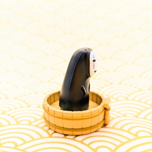 May include: A black and white No Face character from the movie Spirited Away sitting in a small wooden tub. The tub is on a white and gold background.