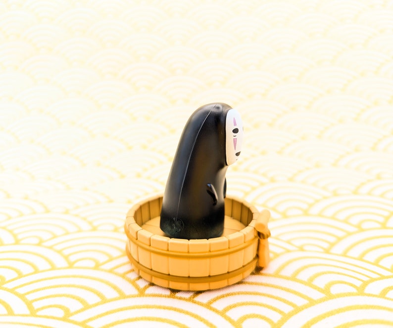 May include: A black and white No Face character from the movie Spirited Away sitting in a small wooden tub. The tub is on a white and gold background.