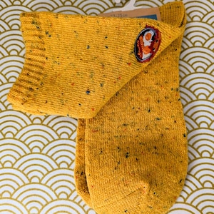 May include: Mustard yellow socks with a speckled pattern and a small embroidered patch. The socks are displayed on a white hanger against a gold and white patterned background. The socks measure 23-25 cm.