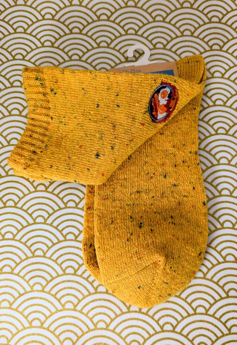 May include: Mustard yellow socks with a speckled pattern and a small embroidered patch. The socks are displayed on a white hanger against a gold and white patterned background. The socks measure 23-25 cm.