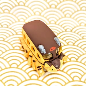 May include: A brown and yellow toy bus with a cat-like face and a brown roof. The bus has a Japanese word "あおい" written on the side. The bus is sitting on a white and gold background.