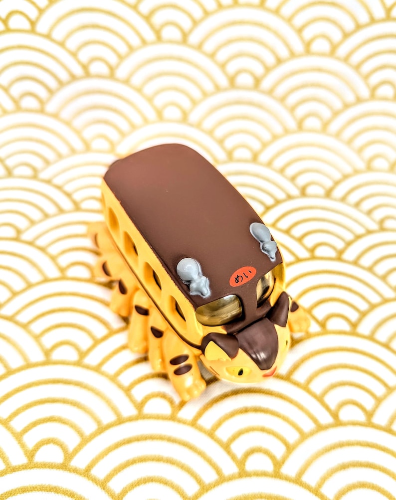 May include: A brown and yellow toy bus with a cat-like face and a brown roof. The bus has a Japanese word "あおい" written on the side. The bus is sitting on a white and gold background.