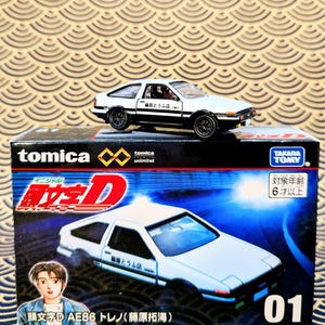 Op de afbeelding: Een wit en zwarte Tomica AE86 modelauto met zwarte wielen staat op zijn doos. De doos heeft de tekst "Initial D" en een illustratie van de auto. De doos heeft ook Japanse tekst en het nummer "01".