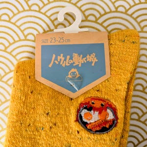 May include: Mustard yellow socks with a patch featuring a cartoon breakfast. The socks have a size tag that reads 23-25 cm. The background is a gold and white wave pattern.