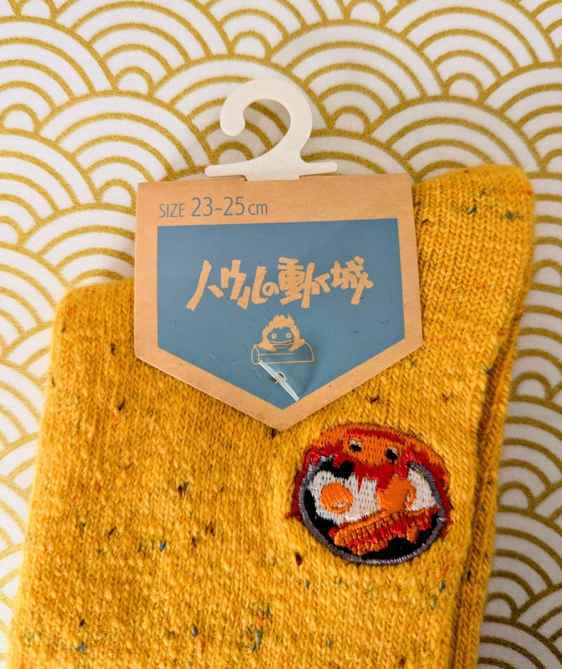 May include: Mustard yellow socks with a patch featuring a cartoon breakfast. The socks have a size tag that reads 23-25 cm. The background is a gold and white wave pattern.