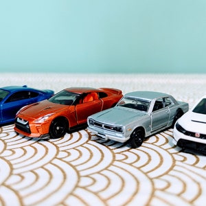 May include: Four miniature toy cars in different colors and styles. A blue car, an orange car, a silver car, and a white car.