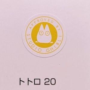 May include: A white sticker with a yellow circle containing the text "APPROVED BY STUDIO GHIBLI" and a cartoon image of Totoro. The text "トロ 20" is printed below the sticker.