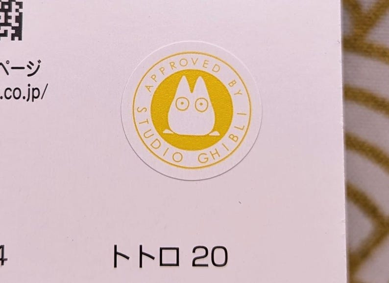 May include: A white sticker with a yellow circle containing the text "APPROVED BY STUDIO GHIBLI" and a cartoon image of Totoro. The text "トロ 20" is printed below the sticker.