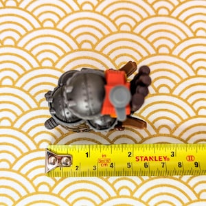 Peut inclure: Petite figurine m&eacute;tallique grise d&eacute;taill&eacute;e avec des accents rouges et bruns, mesurant environ 5 cm de long. La figurine est pos&eacute;e sur une surface &agrave; motifs or et blanc, avec un m&egrave;tre ruban Stanley &agrave; proximit&eacute;.