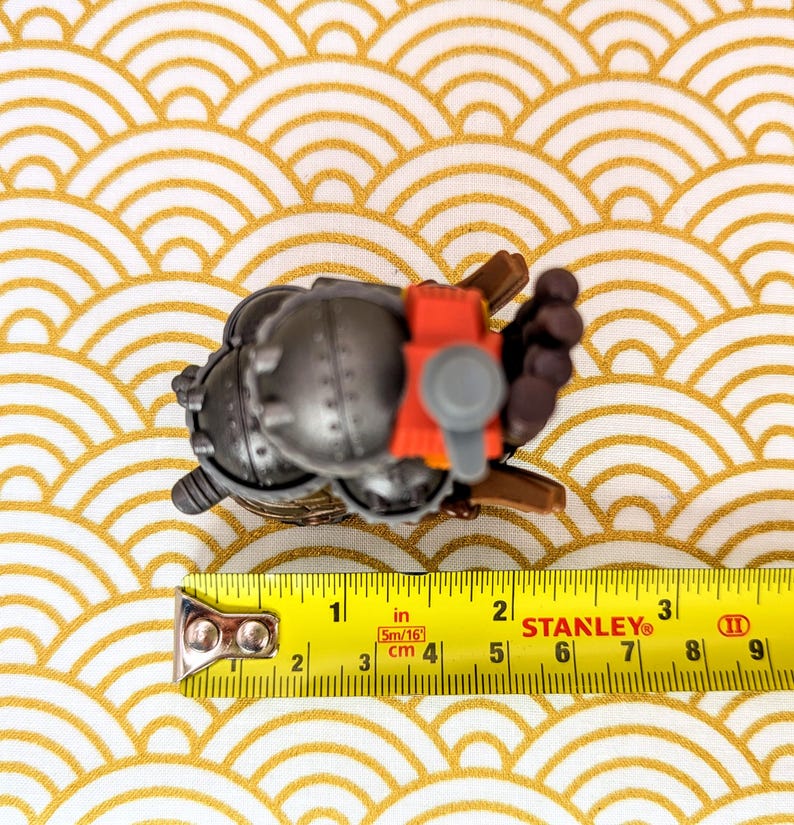 Peut inclure: Petite figurine m&eacute;tallique grise d&eacute;taill&eacute;e avec des accents rouges et bruns, mesurant environ 5 cm de long. La figurine est pos&eacute;e sur une surface &agrave; motifs or et blanc, avec un m&egrave;tre ruban Stanley &agrave; proximit&eacute;.