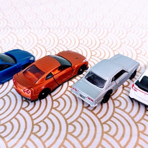 May include: Four miniature toy cars in different colors: blue, orange, silver, and white. The cars are arranged in a row on a white and gold patterned surface.