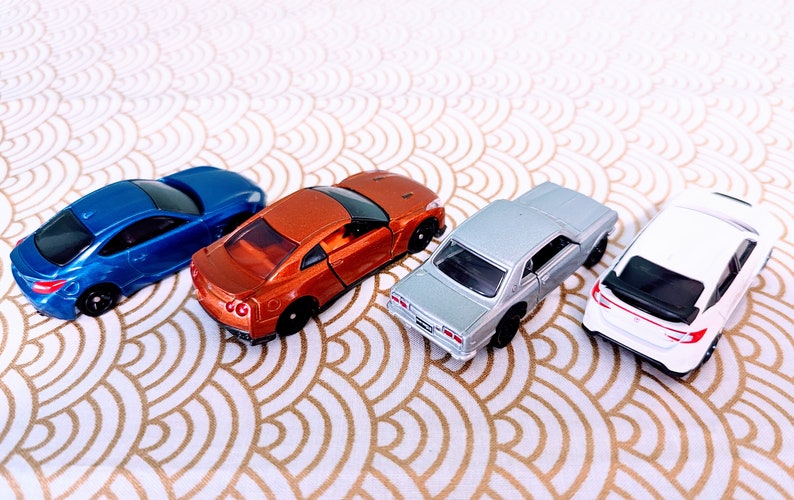May include: Four miniature toy cars in different colors: blue, orange, silver, and white. The cars are arranged in a row on a white and gold patterned surface.