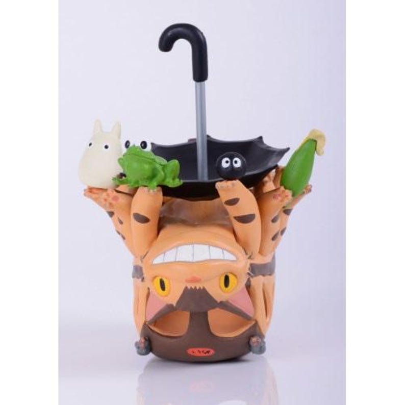 May include: A whimsical figurine featuring a large, orange, bear-like creature holding a black umbrella. A white creature, a green frog, and a green corn cob are perched on the figurine. The umbrella has a black handle.