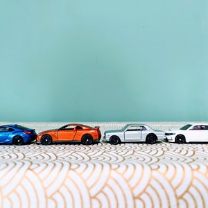 May include: Four miniature toy cars lined up on a white and gold patterned surface. The cars are a blue, orange, silver, and white.