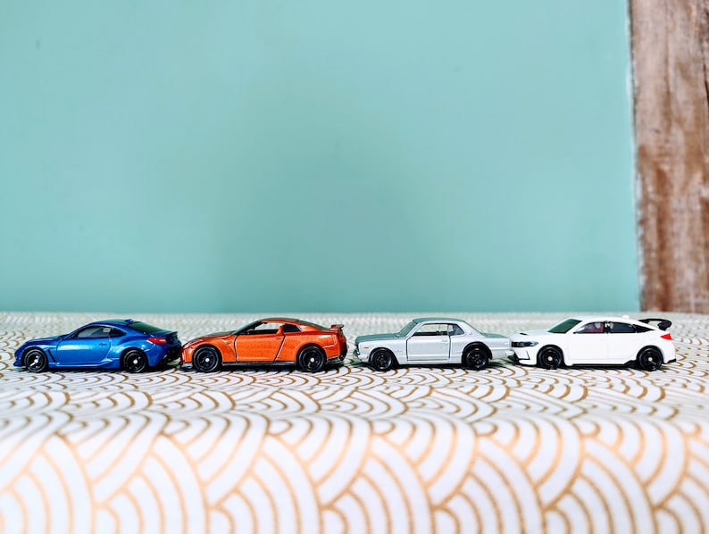 May include: Four miniature toy cars lined up on a white and gold patterned surface. The cars are a blue, orange, silver, and white.