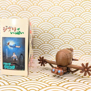 May include: A brown and tan toy airplane with a propeller on each wing. The airplane is sitting on a gold and white patterned surface. The text on the box behind the airplane reads "ジブリ" and "Laputa: Castle in the Sky".