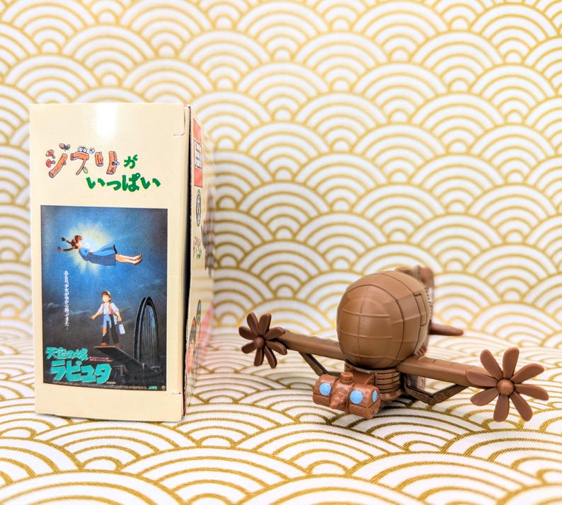 May include: A brown and tan toy airplane with a propeller on each wing. The airplane is sitting on a gold and white patterned surface. The text on the box behind the airplane reads "ジブリ" and "Laputa: Castle in the Sky".