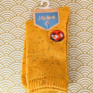 Può includere: Calzini giallo senape con una piccola toppa ricamata con un disegno di granchio. I calzini, taglia 23-25 cm, hanno una trama maculata. Sono appesi a un gancio bianco su uno sfondo a motivi oro e bianco.