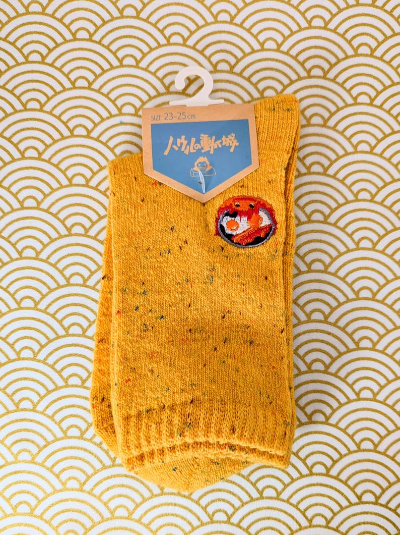 May include: Mustard yellow socks with a small embroidered patch featuring a crab design. The socks are size 23-25 cm and have a speckled texture. The socks are hanging on a white hanger against a gold and white patterned background.