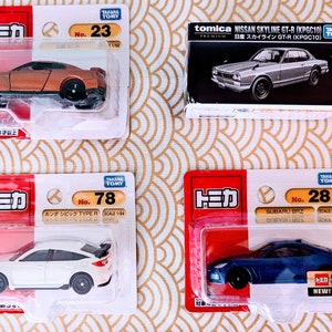 May include: Four toy car models in their packaging. The cars are a gold Nissan GT-R, a white Honda Civic Type R, a blue Subaru BRZ, and a silver Nissan Skyline GT-R. The packaging is white with red and black accents. The cars are all in scale 1/64 or 1/60.