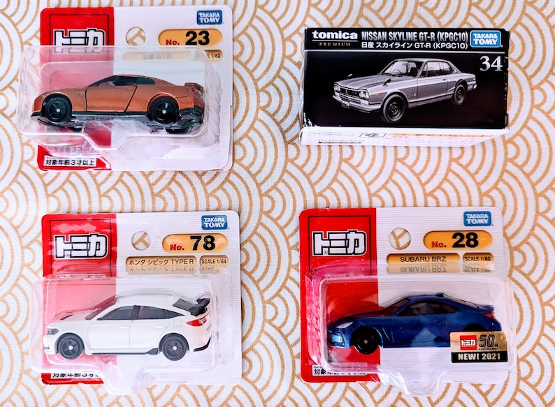 May include: Four toy car models in their packaging. The cars are a gold Nissan GT-R, a white Honda Civic Type R, a blue Subaru BRZ, and a silver Nissan Skyline GT-R. The packaging is white with red and black accents. The cars are all in scale 1/64 or 1/60.