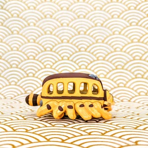 May include: A yellow and brown toy bus with a cat-like face and six legs. The bus has a row of windows and is sitting on a white and gold patterned surface.