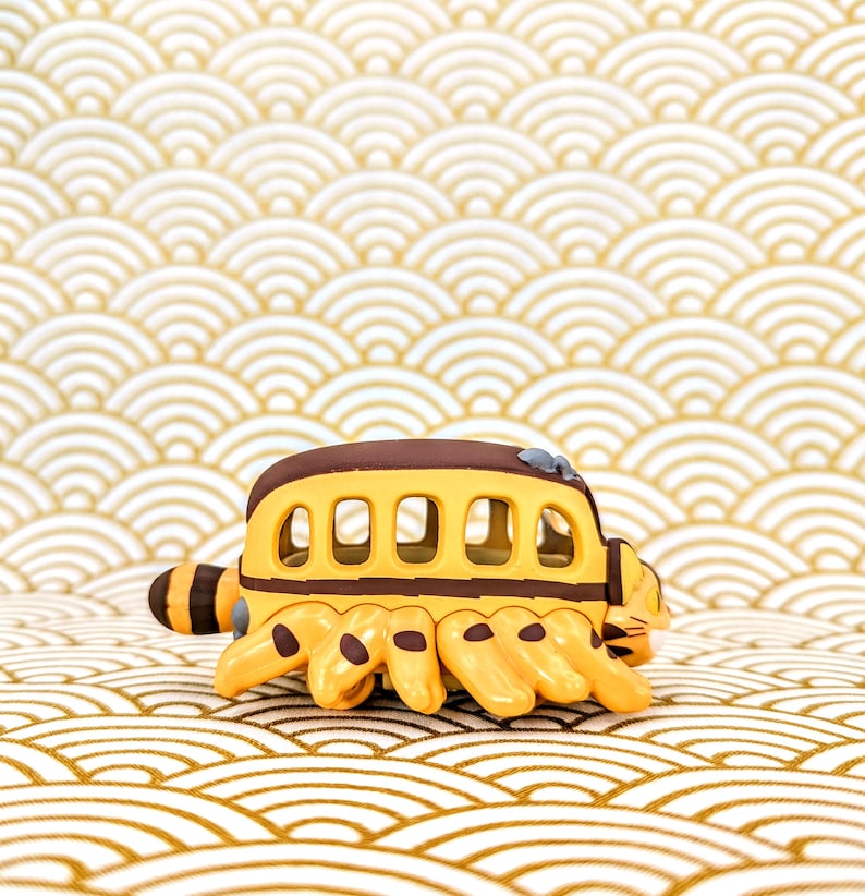 May include: A yellow and brown toy bus with a cat-like face and six legs. The bus has a row of windows and is sitting on a white and gold patterned surface.