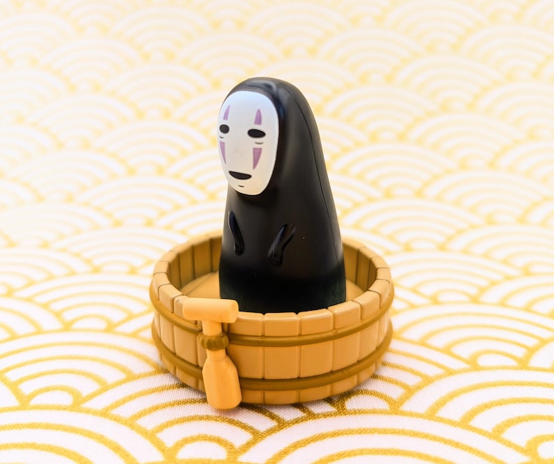 May include: A black and white No Face character from the movie Spirited Away is sitting in a wooden bath tub. The tub is decorated with a light brown wood grain pattern.