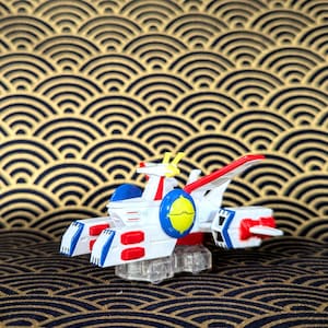 May include: A white, red, blue, and yellow toy robot with a clear plastic base. The robot has a circular blue and yellow emblem on its side and is set against a gold and navy blue patterned background.
