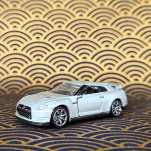 May include: A white model car with a spoiler and silver wheels. The car is set against a background of a gold and navy blue wave pattern.