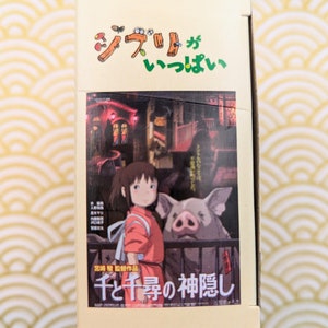 May include: A cardboard box with a colorful illustration of a young girl and a pig. The box has Japanese text that reads "Studio Ghibli" and "Spirited Away".
