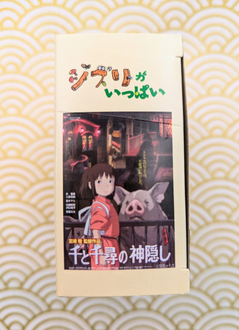 May include: A cardboard box with a colorful illustration of a young girl and a pig. The box has Japanese text that reads "Studio Ghibli" and "Spirited Away".