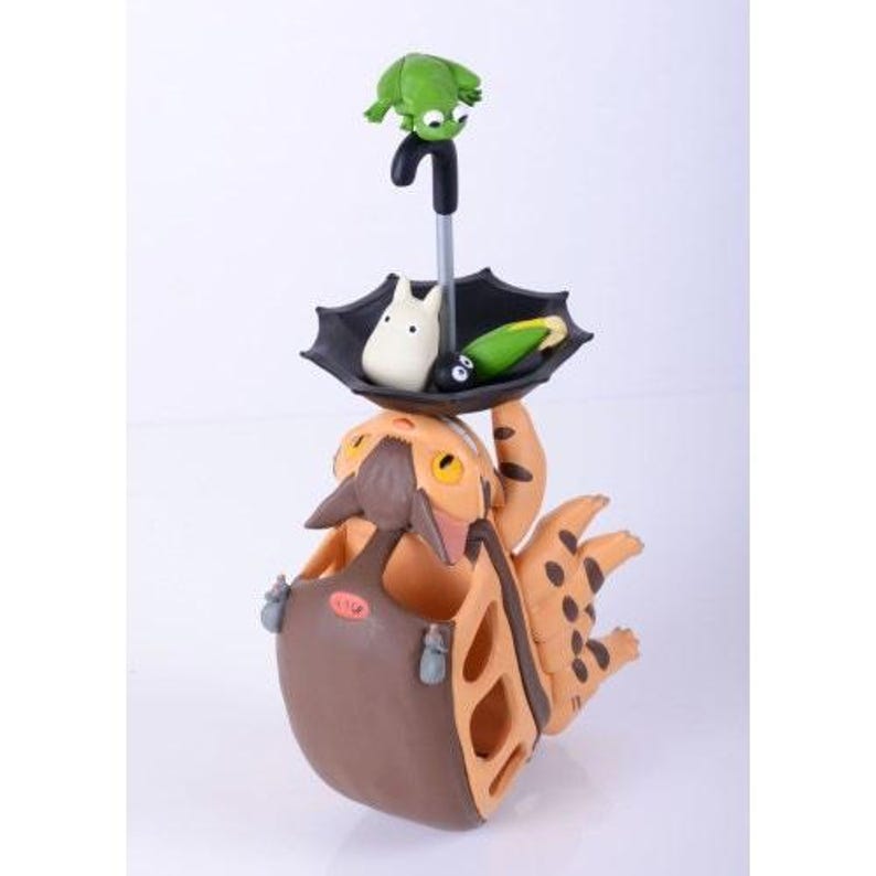 May include: A whimsical figurine featuring a tan and brown creature holding a brown pouch. A black umbrella supports a white figure, a green creature, and a green and yellow object. A green frog sits atop the umbrella. The figurine has a cartoonish style.