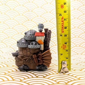 Peut inclure: Figurine de style steampunk marron et gris avec un toit rouge et orange, mesurant environ 7,6 cm de haut. La figurine a plusieurs chemin&eacute;es et une structure en forme d'&eacute;ventail sur le c&ocirc;t&eacute;. Un m&egrave;tre ruban Stanley jaune est &agrave; c&ocirc;t&eacute; de la figurine.