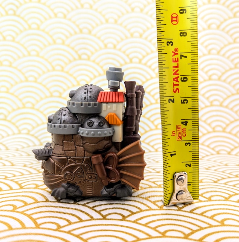 Peut inclure: Figurine de style steampunk marron et gris avec un toit rouge et orange, mesurant environ 7,6 cm de haut. La figurine a plusieurs chemin&eacute;es et une structure en forme d'&eacute;ventail sur le c&ocirc;t&eacute;. Un m&egrave;tre ruban Stanley jaune est &agrave; c&ocirc;t&eacute; de la figurine.