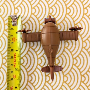 May include: A brown toy airplane with blue eyes and a propeller on each wing. The airplane is about 5 inches long and is sitting on a gold patterned surface.