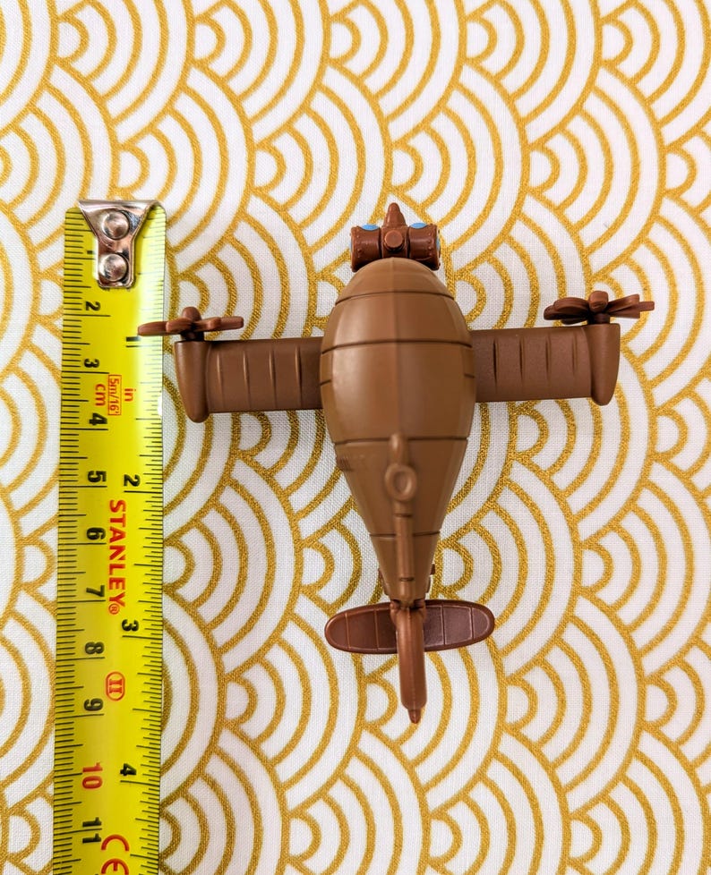 May include: A brown toy airplane with blue eyes and a propeller on each wing. The airplane is about 5 inches long and is sitting on a gold patterned surface.