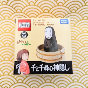 May include: A cardboard box with a colorful illustration of a character from the animated film Spirited Away. The box features the text "Dream Tomica" and "Spirited Away" in Japanese. The box is for a collectible toy figure of No Face from the film.